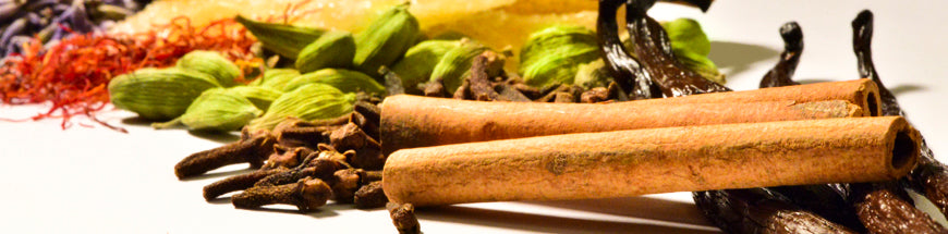 Close-up of assorted spices including cinnamon sticks, cloves, cardamom pods, dried red saffron, vanilla beans, and other colorful spices arranged on a white surface.