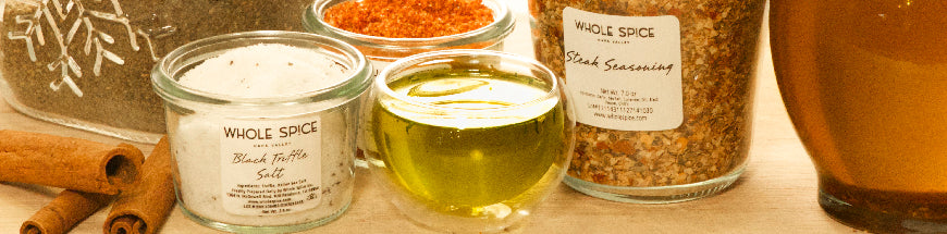 Jars of spices and seasonings, a glass cup with olive oil, cinnamon sticks, and a jar with a snowflake design arranged on a counter in warm lighting.