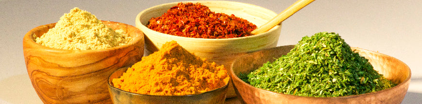 Four wooden bowls filled with colorful powdered spices: yellow, orange, red, and green, with a small spoon resting in the red spice bowl, all arranged on a light surface.