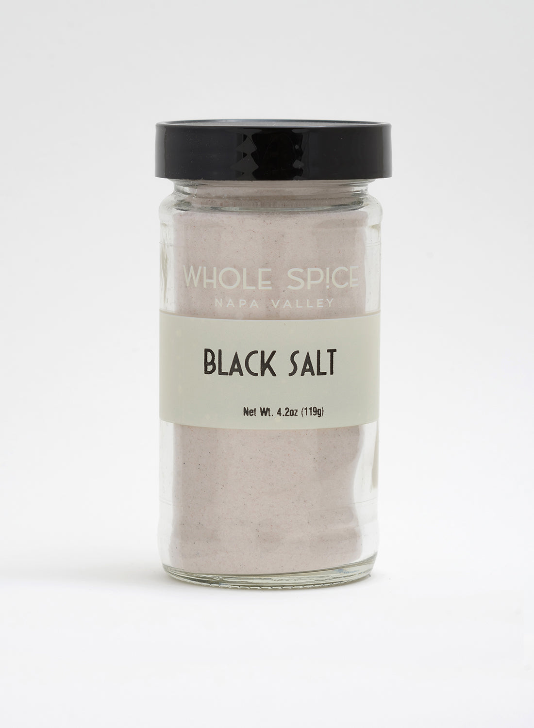 A clear glass jar with a black lid contains finely ground Indian Black Salt (kala namak) by Whole Spice Inc. The label reads “Whole Spice Inc. Black Salt, Net Wt. 4.2oz (119g).” The background is plain white.