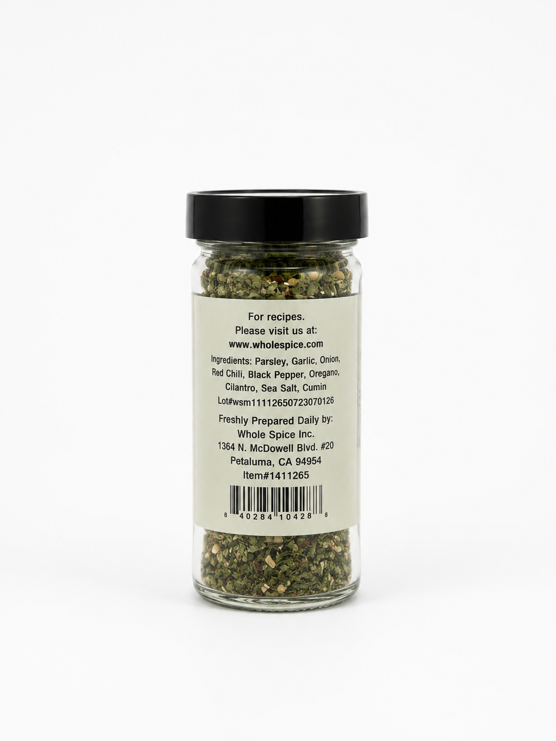 A clear spice jar with a black lid displays a label featuring the ingredients and contact information for Whole Spice Inc., such as their address, website, and lot number. The jar holds Chimichurri Seasoning by Whole Spice Inc., a green and brown herb and spice blend ideal for seasoning grilled meats or bringing authentic South American chimichurri flavor to your recipes.