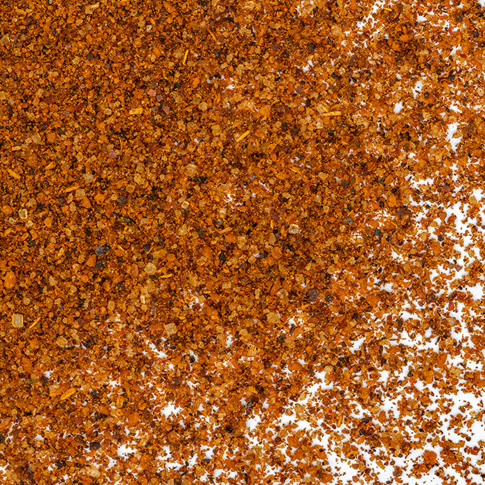 A close-up of Whole Spice Inc. Mole, a reddish-brown Mexican spice blend scattered on a white surface, highlights its coarse, grainy texture with small specks of various sizes.
