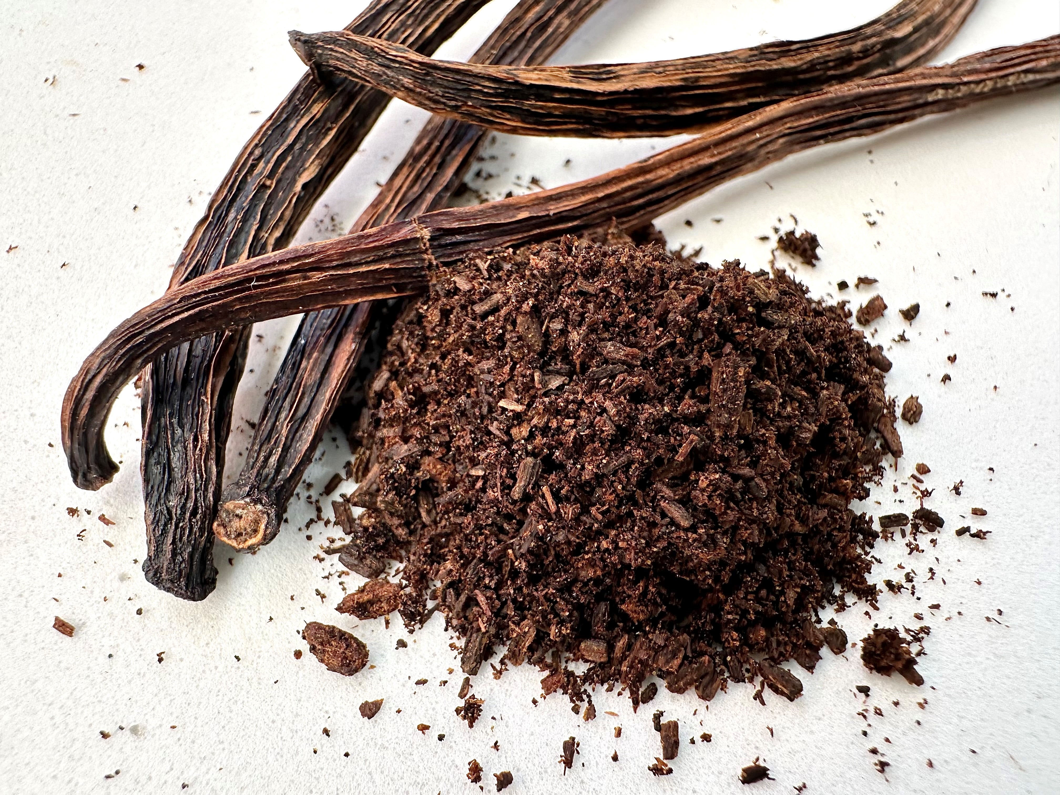 Three whole Whole Spice, Inc. Vanilla Beans for Grinding are placed beside a small heap of dark brown, coarsely ground vanilla bean powder on a white surface.
