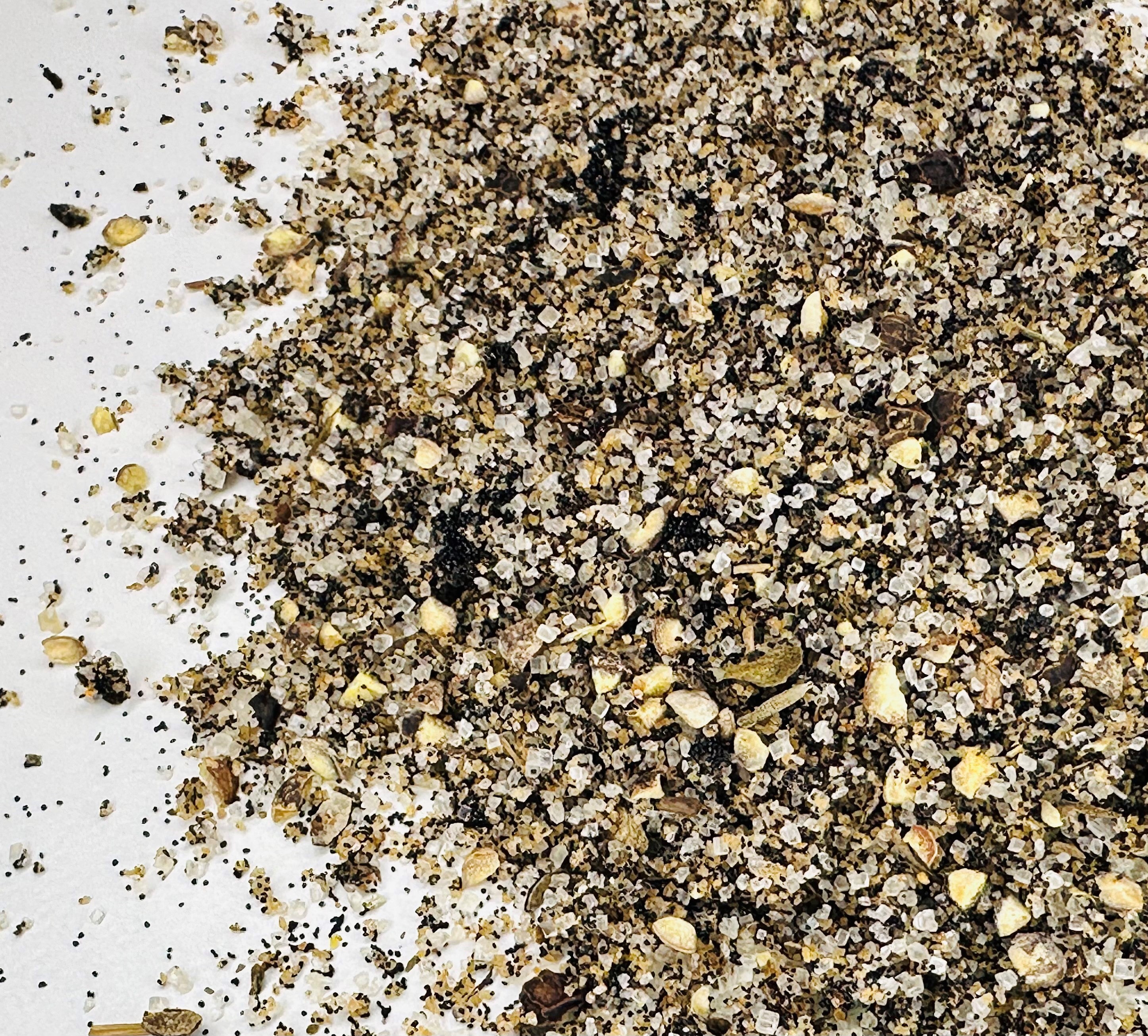 A close-up of Whole Spice Inc.'s Vanilla Pepper Rub—a coarse gourmet blend of black pepper, salt, and spices scattered on a white surface—perfect as a meat rub.