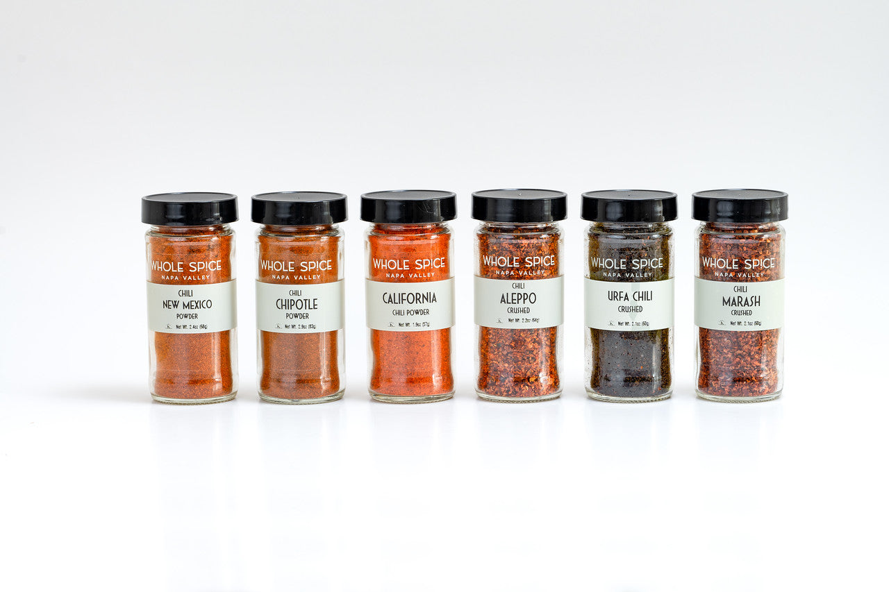 Six glass jars with black lids from Whole Spice Inc.’s Chili Gift Set contain New Mexico, Chipotle, California, Aleppo, Urfa Chili, and Marash—an ideal gift for any spice lover.
