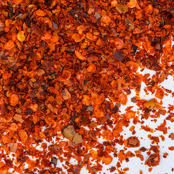 Close-up of Whole Spice Inc. Aleppo Chili Crushed, scattered on a white surface, reveals vibrant red, orange, and brown flakes with yellow seeds, commonly used to add Mediterranean flavor.