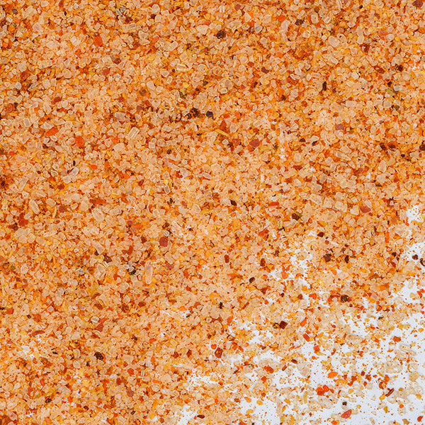 Close-up of Whole Spice Inc. All Purpose Salt Seasoning—an orange and red spice blend with a coarse, granular texture—densely scattered across a white surface.