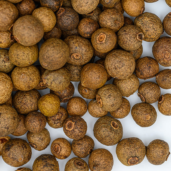 A close-up of Whole Spice Inc. Allspice Whole berries scattered on a white surface, showcasing their round shape and rough brown texture.