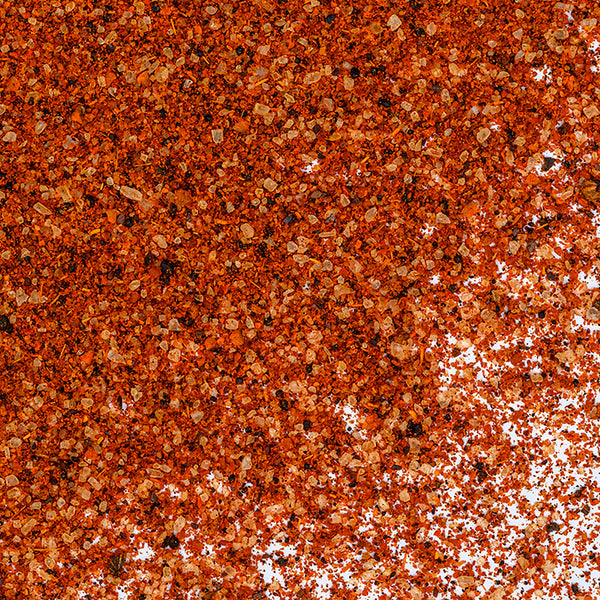 Close-up of Whole Spice Inc. BBQ Rub—coarse red and orange granules scattered across the surface, with some white background visible at the lower right where the spice rub thins out.