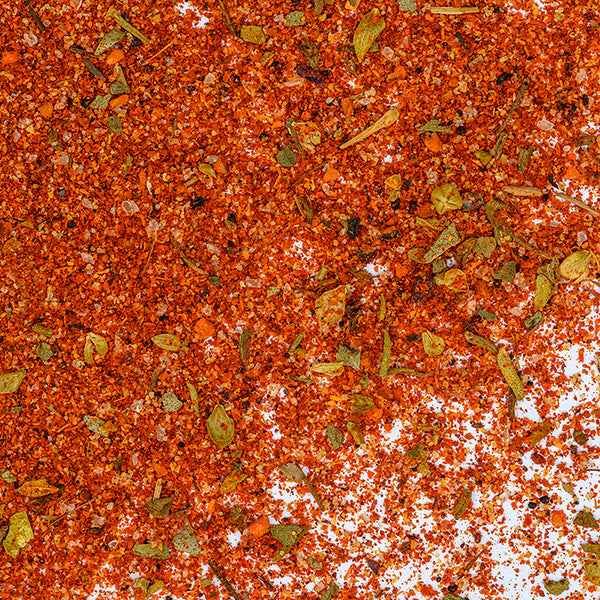 A close-up of Whole Spice Inc. Blackened Seasoning shows a vibrant, textured mix of spices, dried herbs, and seeds scattered across a white surface.