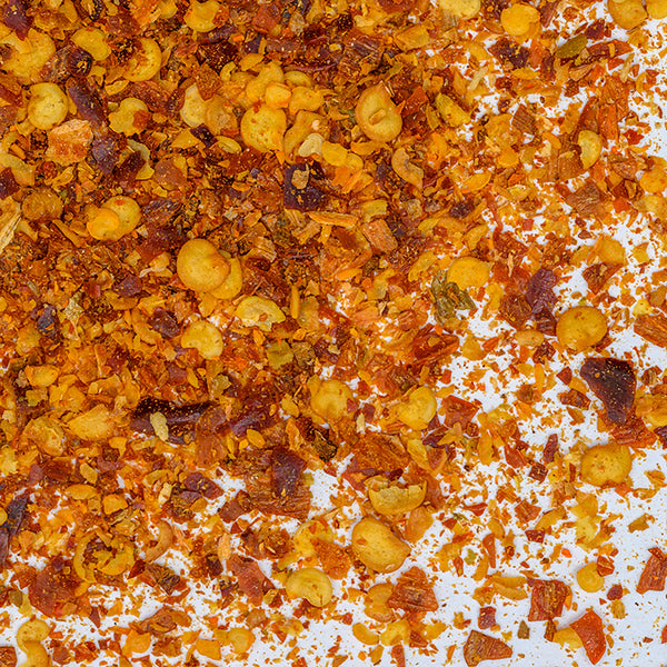 Close-up of Whole Spice Inc. Calabrian Peperoncino Chili Flakes scattered on a white surface, revealing a vibrant mix of red, orange, and yellow pieces in various sizes and textures.