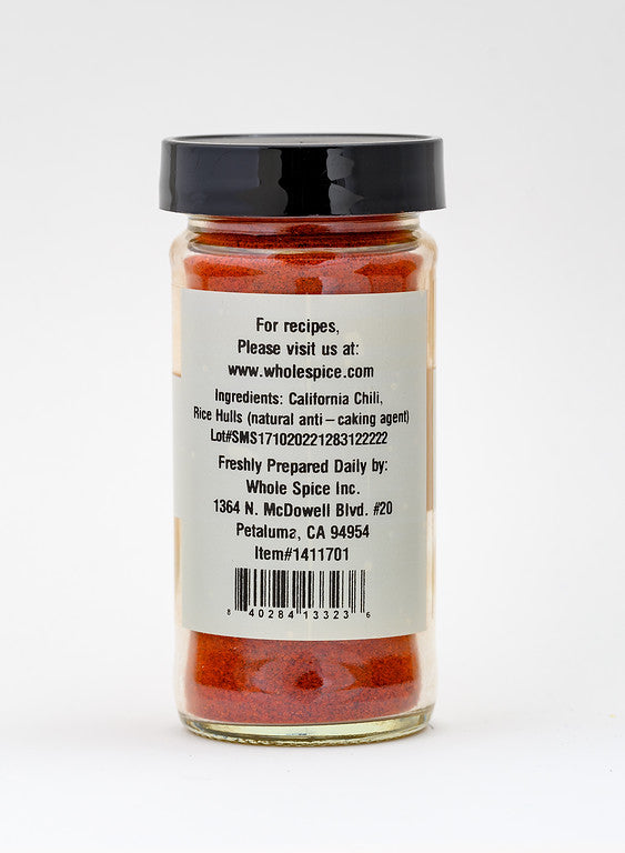 A clear glass spice jar with a black lid contains California Paprika. The back label lists ingredients, Whole Spice, Inc. address, website, lot number, and item number on a gray background.