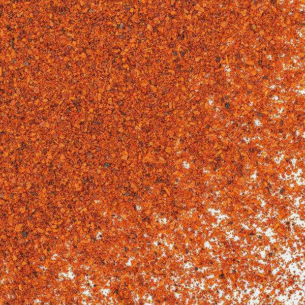 A close-up of Whole Spice Inc. Chili Seasoning—finely ground red-orange powder scattered on a white surface, densely layered in the upper left and thinning toward the lower right—ideal for adding bold flavor to your favorite chili recipes.
