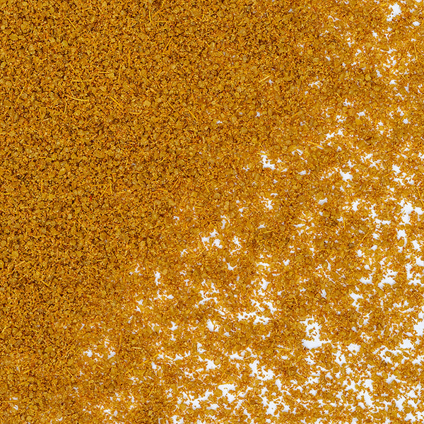 Close-up of golden brown Whole Spice Inc. Cumin Powder granules scattered on a white background, densely packed in the top left and more spread out toward the bottom right.