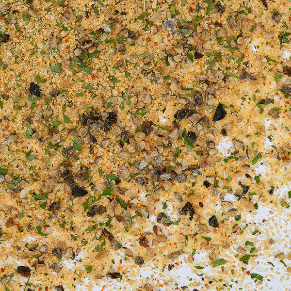 A close-up of Whole Spice Inc.’s Garlic Pepper Seasoning, with dried herbs, coarse black pepper, green flakes, and fine yellowish granules on a white background—ideal as a meat seasoning.