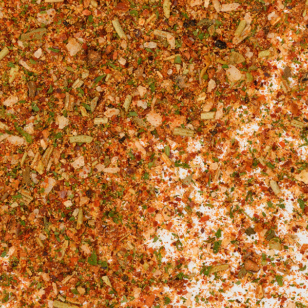 A close-up of Whole Spice Inc. Mediterranean Roasting Rub shows visible herbs, red pepper flakes, green and orange seasonings, and coarse kosher salt granules scattered on a white background.