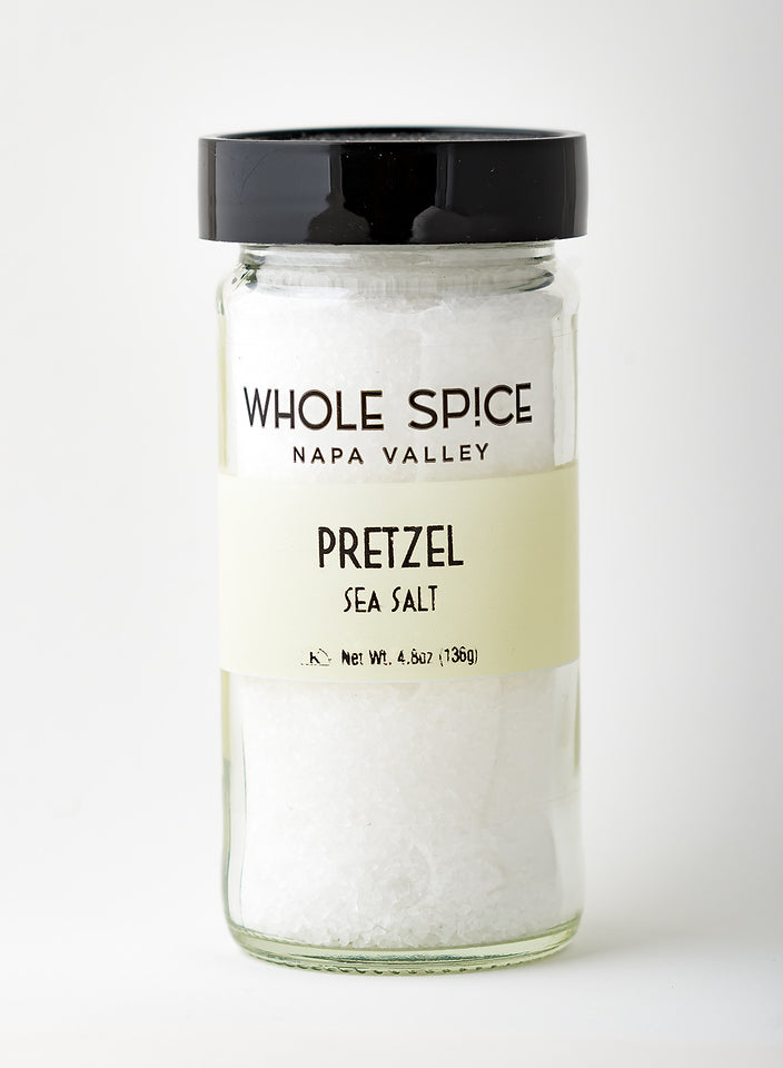 A glass jar of Whole Spice Inc. Pretzel Sea Salt with a black lid, filled with coarse white sea salt, features a cream-colored label showing the product name and weight.