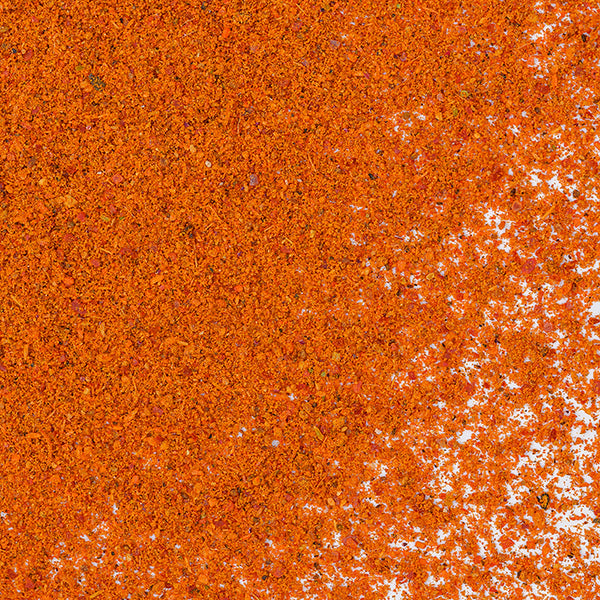 A close-up of Whole Spice Inc. Ras el Hanout Ground shows a pile of orange-red powder with fine and coarse granules scattered on a white surface, reflecting the vibrant colors and textures of this Moroccan spice blend.