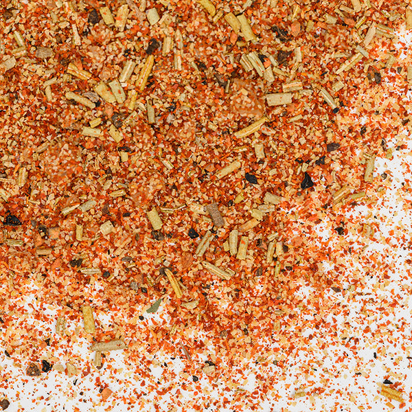 A close-up of Whole Spice Inc.'s Roasted Chicken Blend Organic, showcasing vibrant red, orange, yellow, and brown seasoning with dried herbs and French gray salt scattered on a white surface.