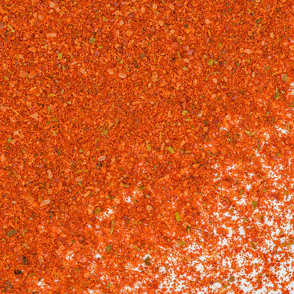 A close-up of Whole Spice Inc. Tandoori Spice Rub shows its vibrant orange-red color scattered unevenly across a white surface, with both densely covered and lightly dotted areas.