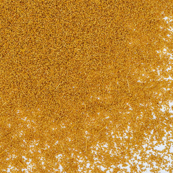 Close-up of a pile of golden-brown granules, identified as Whole Spice Inc. Cumin Powder Toasted, scattered unevenly on a white background.