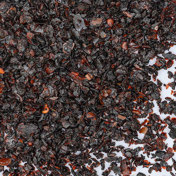 Close-up of Whole Spice Inc. Urfa Chili Crushed flakes, dark brown to reddish-black, scattered on a white background; their coarse, irregular texture and small fragments resemble crushed tea leaves.