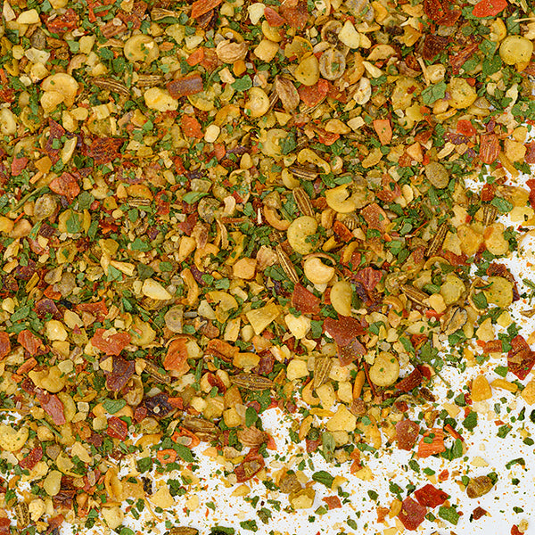 A close-up of Whole Spice Inc. Zhug Spice, a vibrant Yemenite blend with visible dried herbs, red pepper flakes, minced garlic, and assorted seasonings scattered on a white background.