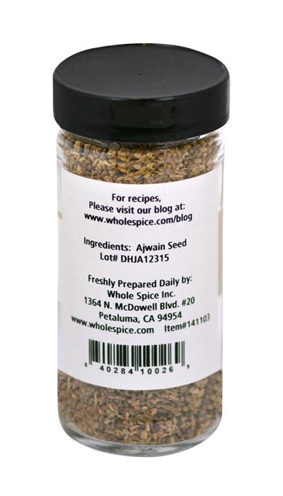 A clear spice jar with a black lid from Whole Spice Inc. contains Ajwain Seed Whole. The label lists ajwain seed, lot number, company address, website, barcode, and a note to visit the blog for recipes.