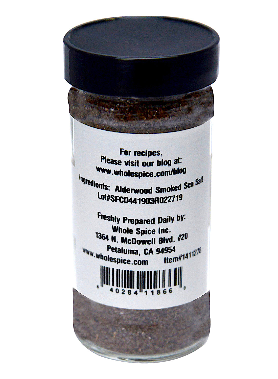 A clear spice jar with a black lid, labeled Alderwood Smoked Sea Salt by Whole Spice Inc., features ingredient info, manufacturer's contact details, a recipes website, and packaging info from Petaluma, California.