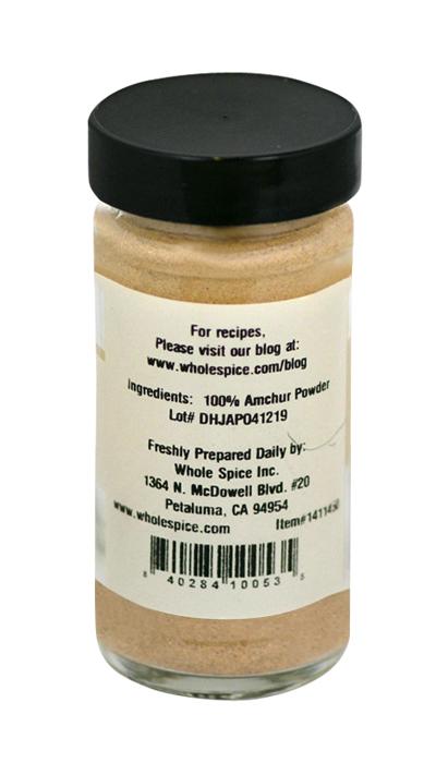 A clear spice jar with a black lid, labeled on the back. The label reads "Amchur Powder" by Whole Spice Inc., lists 100% amchur powder, company info, address, website for recipes, barcode, and lot number.