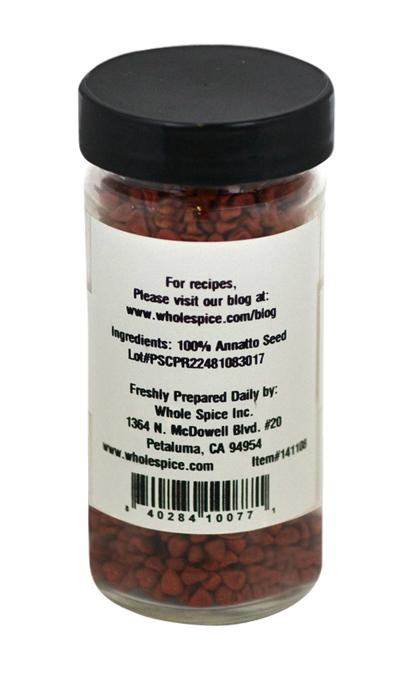 A clear spice jar from Whole Spice Inc., labeled "Annatto Seeds," has a black lid and is filled with reddish-orange achiote seeds, a natural coloring. The label displays ingredients, company details, and a barcode.