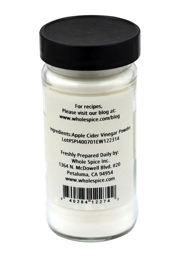 A clear spice jar with a black lid holds Whole Spice Inc. Apple Cider Vinegar Powder, a tangy white powder. The label features company info, address, website, and barcode—ideal for seasoning blends.