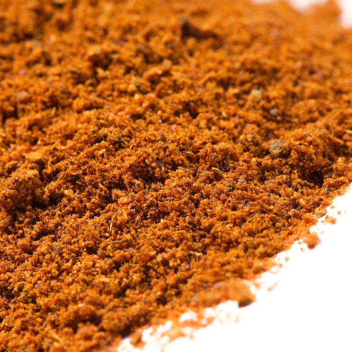 A close-up of finely ground Baharat Spice by Whole Spice Inc., an aromatic reddish-brown Arabic blend popular in Gulf States cuisine, scattered on a white surface.