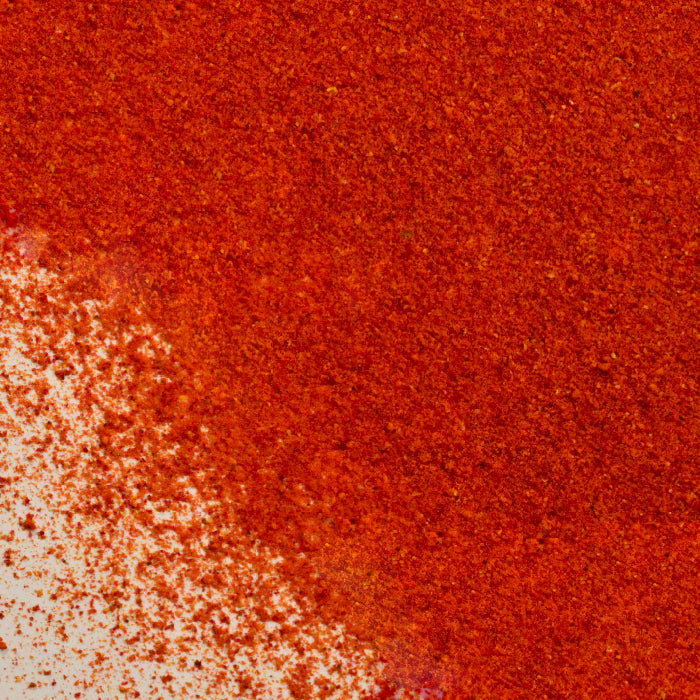 Close-up of Roasted Red Bell Pepper Powder by Whole Spice Inc., its vibrant red-orange granules scattered on a white surface, creating a textured, powdery look.