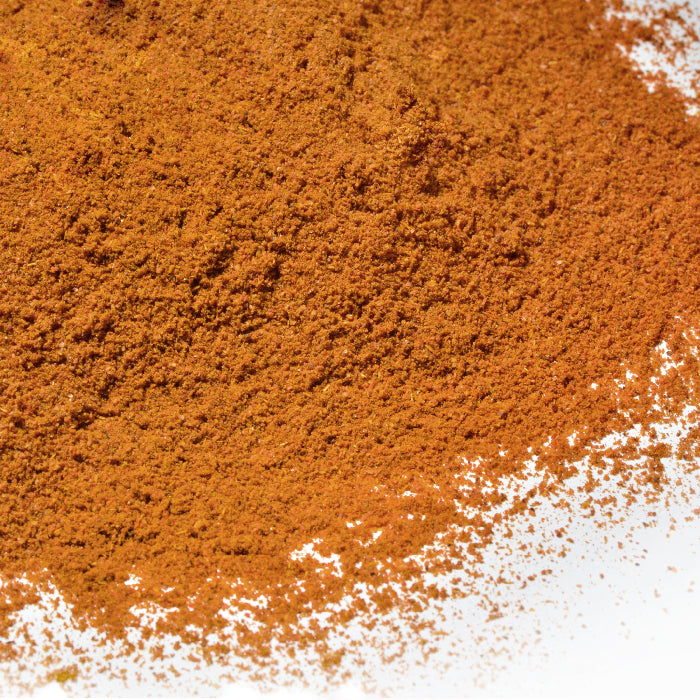 A close-up of Whole Spice Inc. Bird's Eye Chili Powder, a fine, reddish-brown spice, is scattered on a white surface.
