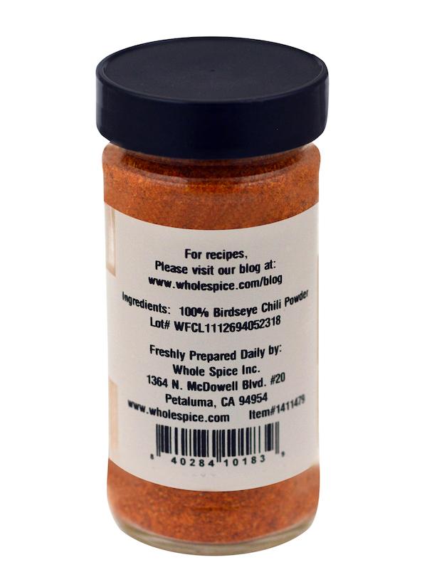 A clear spice jar with a black lid holds orange-red Bird's Eye Chili Powder by Whole Spice Inc. The label shows 100% chili powder, authentic peppers, manufacturer and website info, plus a barcode at the bottom.