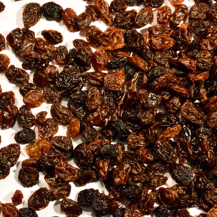 A close-up of Whole Spice Inc. Black Raisins, also known as dried black grapes, scattered on a white surface.