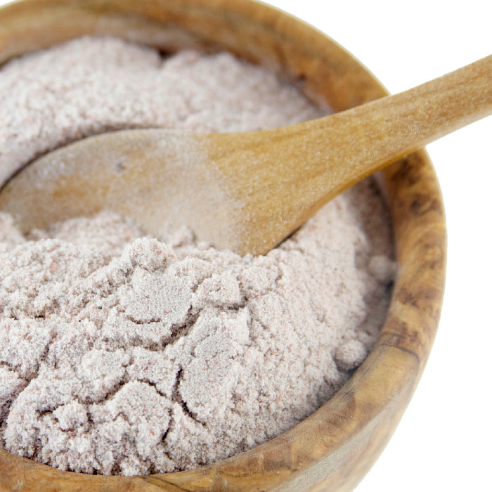 A wooden bowl of light purple Black Salt (kala Namak) from Whole Spice Inc., with a wooden spoon partially buried in the salt, set against a white background.