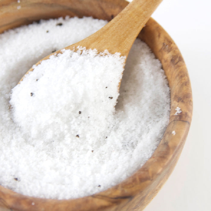 A wooden bowl filled with Whole Spice Inc. Black Truffle Salt, coarse white crystals flecked with black specks, and a wooden spoon scooping up some of the gourmet salt.