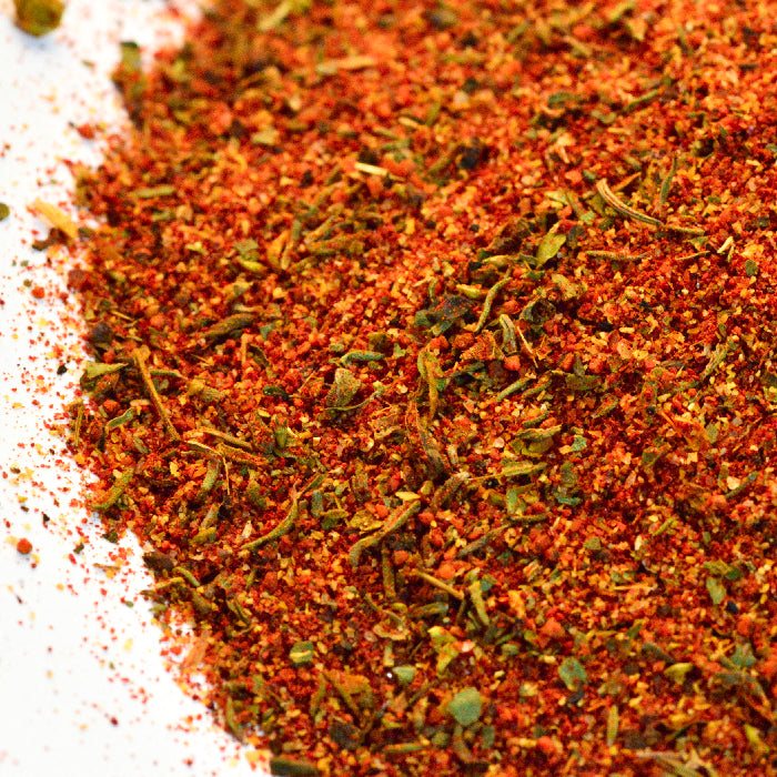 A close-up of Whole Spice Inc. Blackened Seasoning displays a vibrant mix of ground and leafy dried herbs and spices in red, orange, and green flecks on a white surface.