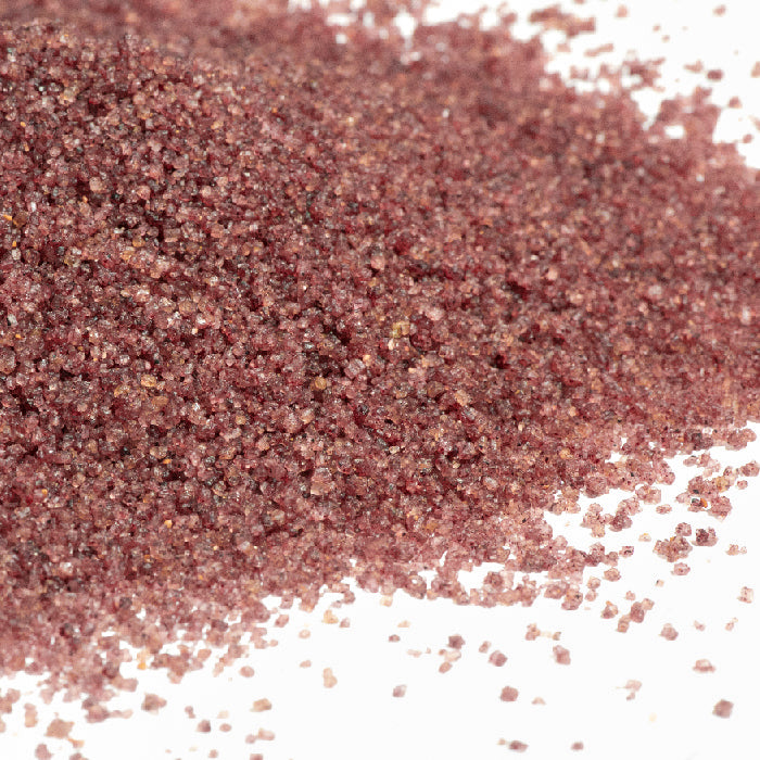 A close-up of coarse, reddish-brown sumac spice grains scattered on a white surface, accented by Whole Spice Inc.'s Wild Blueberry Sugar for a subtle hint of color and sweetness.