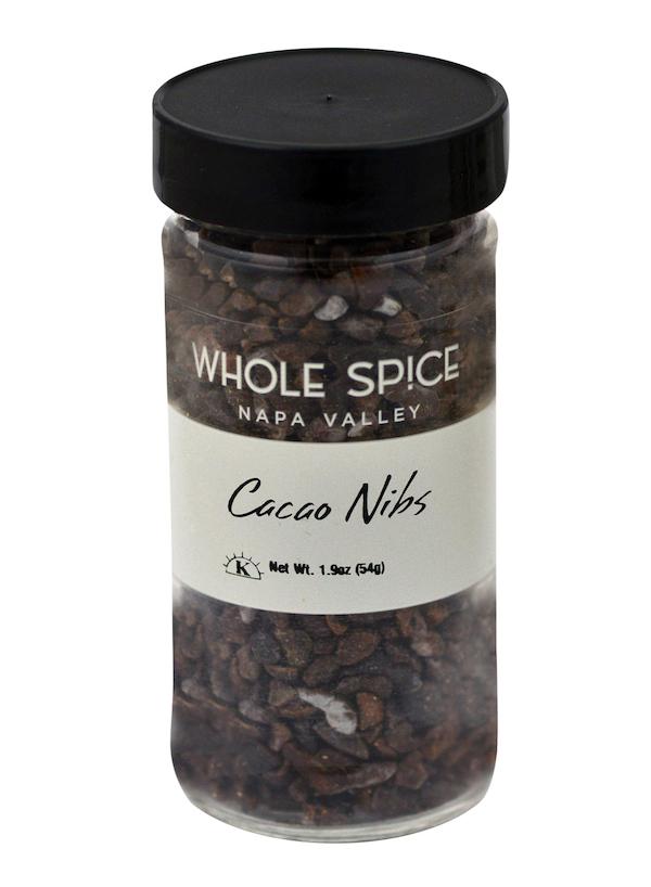 A clear plastic jar with a black lid holds small dark brown cacao nibs from Whole Spice Inc. The label reads "Cacao Nibs" and shows a net weight of 1.9 oz (54g), indicating the product is made from roasted cocoa beans.