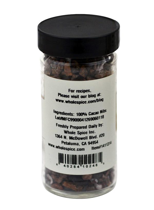 A clear spice jar with a black lid holds Whole Spice Inc.'s Cacao Nibs made from roasted cocoa beans. The white label features ingredients, company address, barcode, “100% Cacao Nibs,” and the Whole Spice Inc. website.