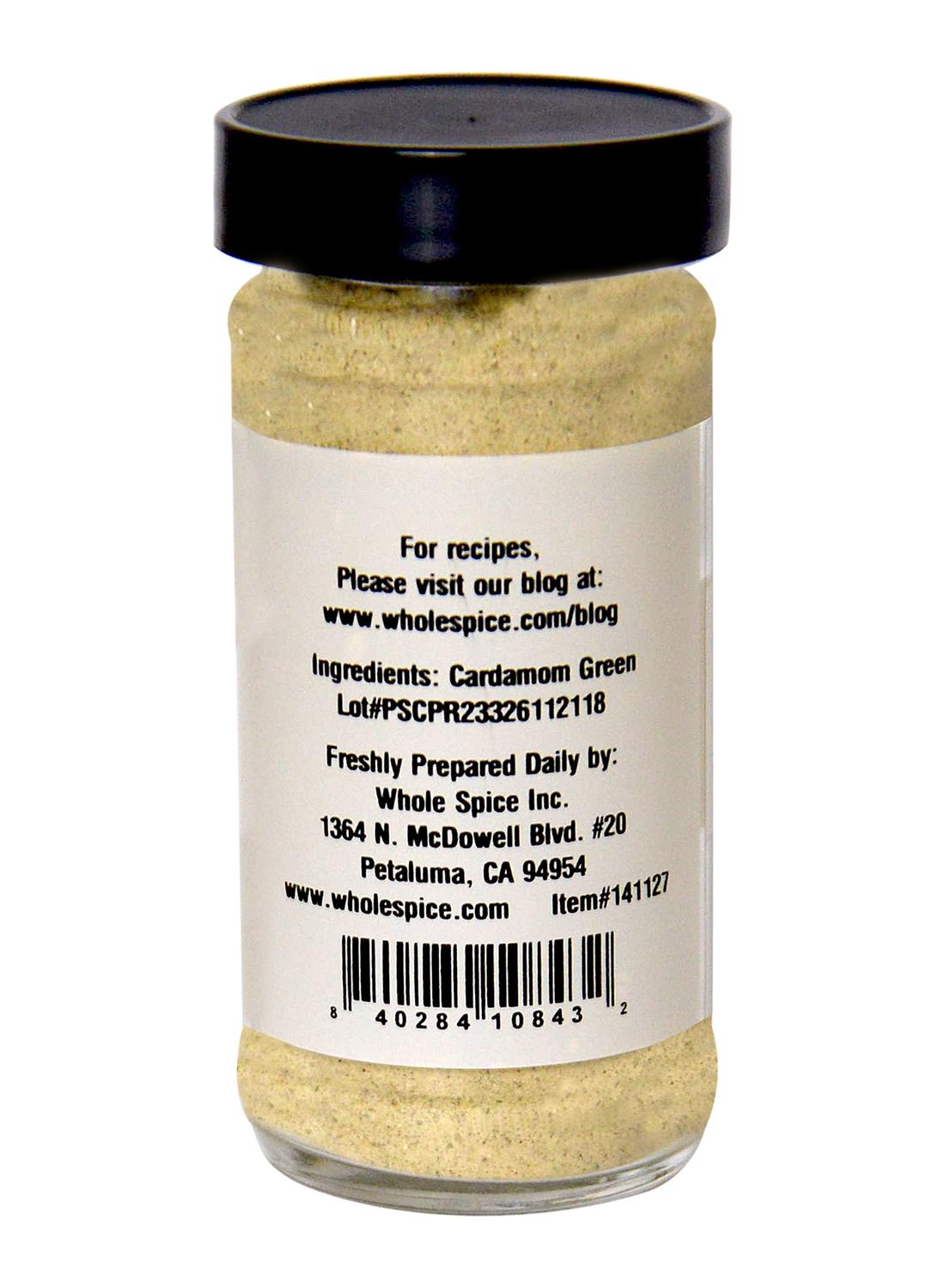 A clear spice jar with a black lid contains light beige Green Cardamom Powder by Whole Spice Inc. The label lists ingredients as Green Cardamom and includes company info, lot number, barcode, and a recipe website.