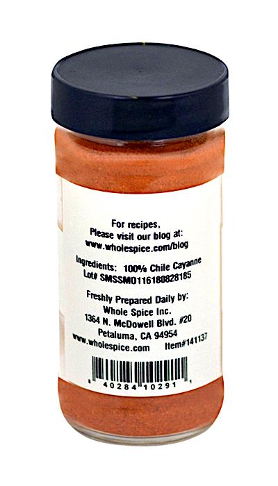 A clear spice jar with a black lid is labeled on the back: Cayenne Chili Powder by Whole Spice Inc., made from 100% cayenne pepper. The label lists contact info, website for recipes, and a barcode at the bottom.