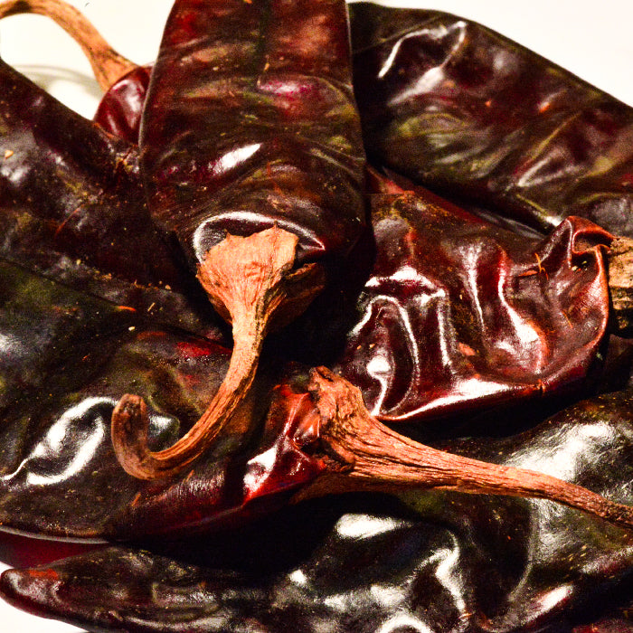 Close-up of Guajillo Chili Whole by Whole Spice Inc.—dried dark red peppers with wrinkled, glossy skins and long, curved stems piled on a light surface—ideal for making mole sauce.