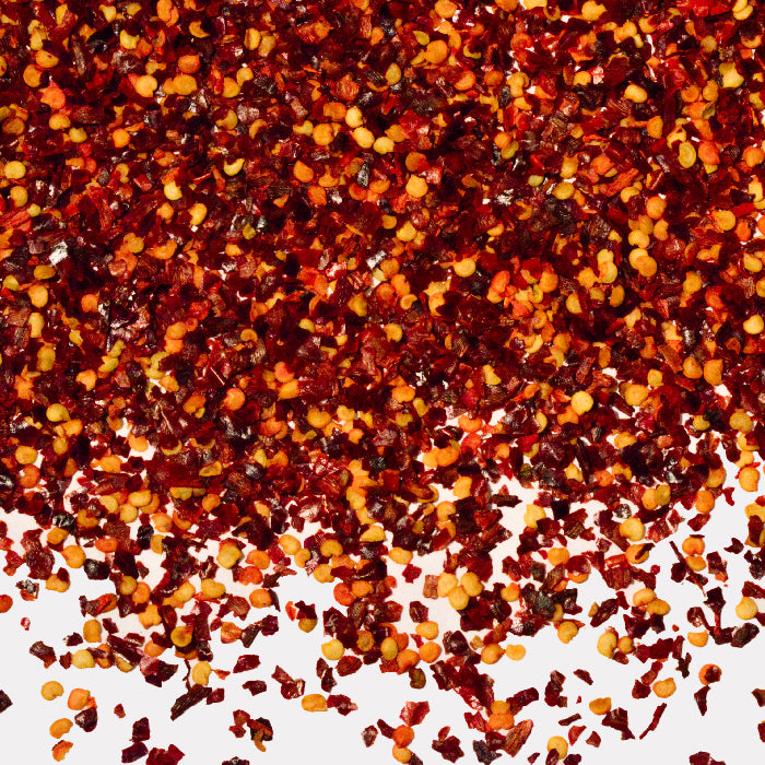 Chili Pepper Flakes by Whole Spice Inc. are shown scattered on a white background, highlighting their vibrant red and orange hues along with visible small seed pieces.