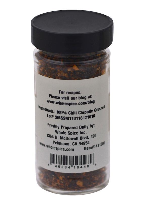A clear jar of Whole Spice Inc. Chipotle Chili Crushed with a black lid contains 100% smoked jalapeño peppers. The label also features distributor details and a barcode.