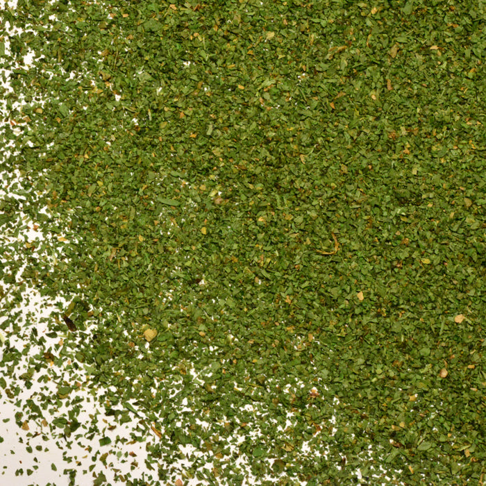 A close-up of dried, chopped cilantro leaves from Whole Spice Inc. scattered on a white surface, showcasing their fine texture and vibrant green color—an essential culinary herb often paired with spicy dishes.