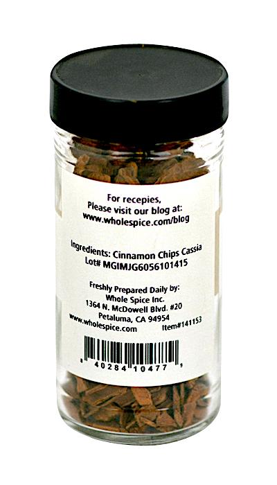 A clear glass jar with a black lid holds premium Cinnamon Chips Cassia by Whole Spice Inc. The label lists ingredients, company details, lot number, contact info, and a barcode at the bottom.